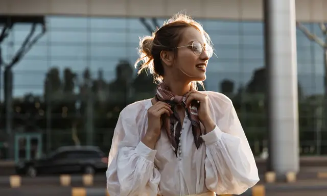 Uśmiechnięta kobieta w okularach poprawia apaszkę na szyi. Pokazuje, jak zawiązać apaszkę, tworząc stylowy look.
