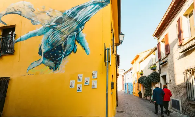 Un murale di balene blu su un muro giallo a San Giuliano Rimini. Una coppia passeggia lungo la strada acciottolata.