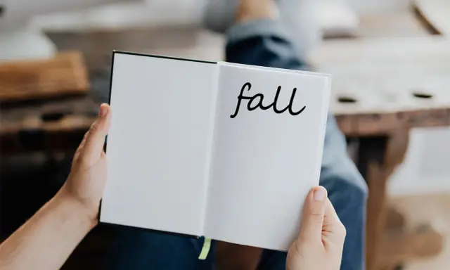 Otwarta książka z napisem "fall" na prawej stronie. Osoba w dżinsach trzyma ją, przygotowując się do jesiennych zapisków.