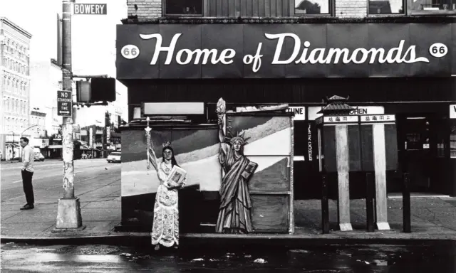 Corky Lee photos: A person in ornate robes stands beside a Statue of Liberty cutout on a wet street, with "Home of Diamonds" sign above.