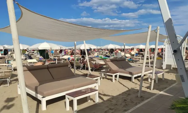 Un lido a Riccione con lettini doppi e ombrelloni bianchi sotto un cielo azzurro e nuvoloso.