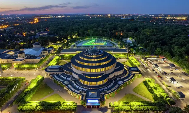Wrocław Hala Stulecia nocą, z podświetloną fontanną i otaczającym parkiem.