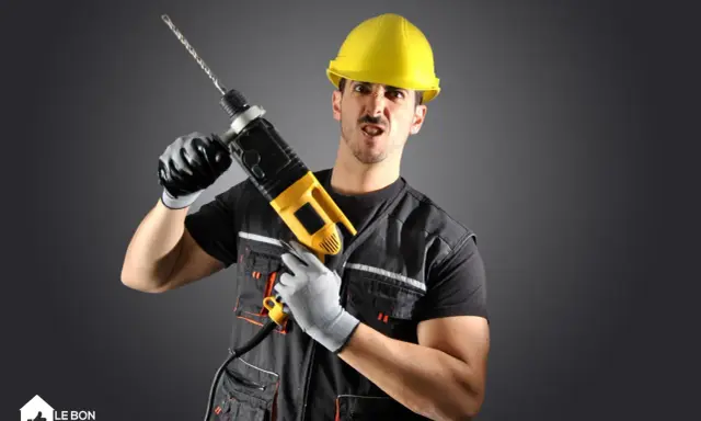 Un homme en casque jaune, gants et tenue de travail tient une perceuse. Il a une expression faciale exagérée, comme un architecte ou un constructeur prêt à relever un défi.