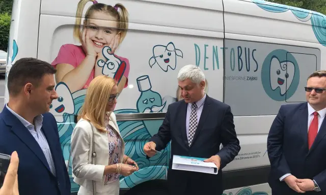 Ludzie stojący przed białym Dentobusem z grafikami uśmiechniętych zębów i dziewczynki.