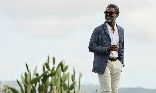 Man in stylish beach wedding attire, holding a drink, with cacti and a hazy sky.