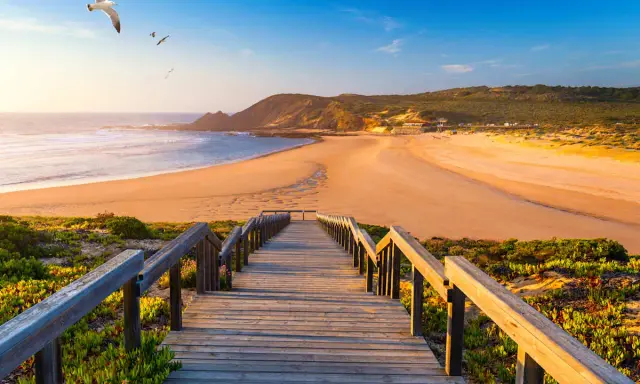 Holzsteg führt zu einem goldenen Strand in Aljezur, Portugal. Möwen kreisen über dem Meer unter blauem Himmel.
