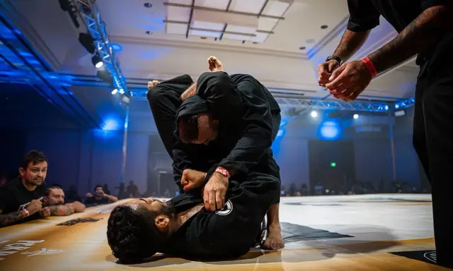 Dwóch mężczyzn w czarnych kimonach walczy w parterze. Intensywna walka jiu jitsu, z widownią w tle.