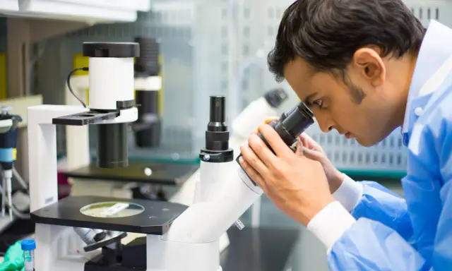 Mężczyzna w niebieskim fartuchu laboratoryjnym patrzy przez mikroskop.