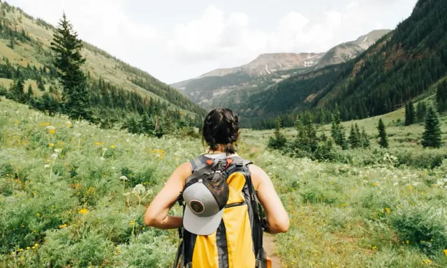 Kobieta z plecakiem wyrusza na trekking przez kwiecistą dolinę, podziwiając górskie krajobrazy.