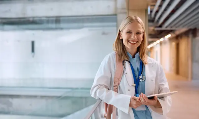 Uśmiechnięta studentka medycyny w białym kitlu, z plecakiem i stetoskopem, trzyma tablet. Zastanawia się, gdzie najłatwiej dostać się na medycynę.