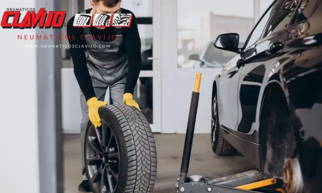 Mecánico con guantes amarillos sosteniendo una rueda, listo para cambiar neumáticos. El coche está en un taller.