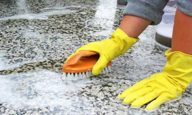 Ręce w żółtych rękawiczkach szorują szczotką płomieniowany granit, usuwając brud i pianę.