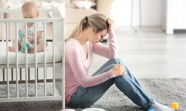 Zmartwiona mama siedzi obok łóżeczka, zastanawiając się, depresja poporodowa ile trwa. Dziecko bawi się w łóżeczku.