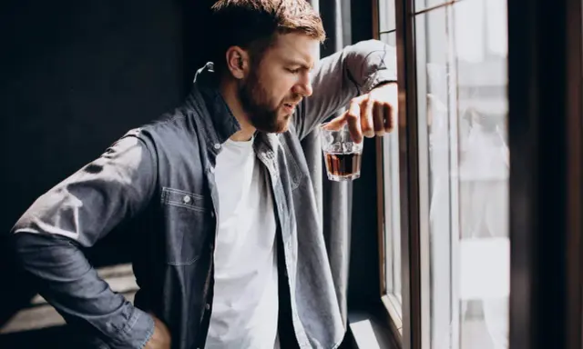 Mężczyzna zamyślony przy oknie, trzymający szklankę z bursztynowym płynem. Czy to jeden z ukryty alkoholizm objawy?