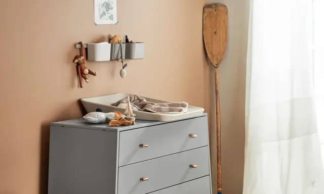 Eine Wickelkommode organisieren: Spielzeug, Handtücher und Pflegeutensilien sind ordentlich verstaut. Ein Holzpaddel lehnt an der Wand.