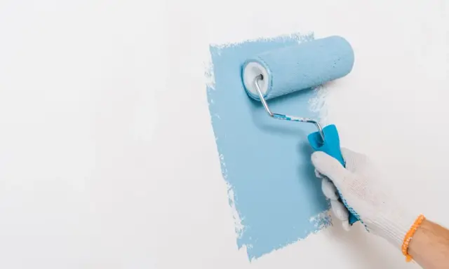 Ręka w białej rękawiczce maluje ścianę na niebiesko wałkiem. Widać nierówności, co może sugerować wady farby ceramicznej.