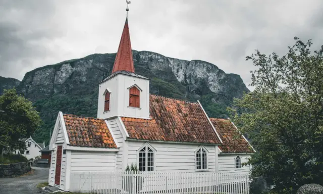 Biały kościółek z czerwoną wieżą i dachem, otoczony białym płotkiem, na tle surowych gór. Symbol norweskiej religii i spokoju.