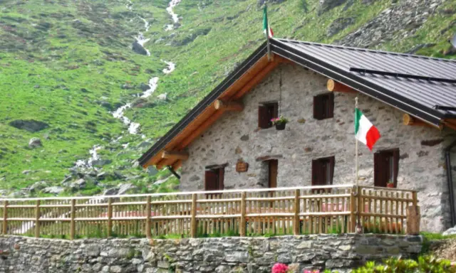 Rifugio in pietra con bandiera italiana, circondato da verdi pendii e un ruscello. Un'oasi accogliente tra i rifugi Val Camonica facili da raggiungere.