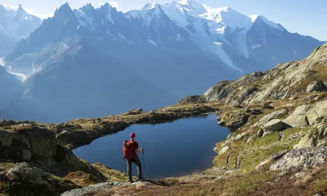 Widok na wejście na Mont Blanc. Turysta z plecakiem i kijkami stoi nad górskim jeziorem, podziwiając ośnieżone szczyty.