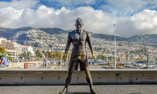 Bronzestatue von Cristiano Ronaldo am Hafen von Funchal, Madeira. Hinter ihm erstreckt sich die Stadt bis zu den Bergen. Ein Denkmal für den weltberühmten Fußballer, das man fast wie am **cristiano ronaldo flughafen** erwarten könnte.