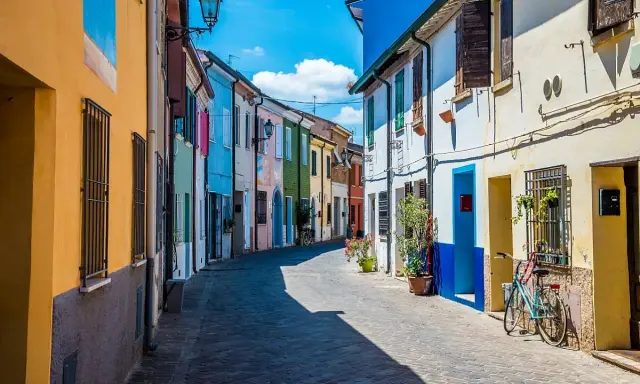 Un vicolo colorato a San Giuliano, con case dipinte di giallo, blu, verde e rosa. Una bicicletta azzurra è parcheggiata davanti a una casa.