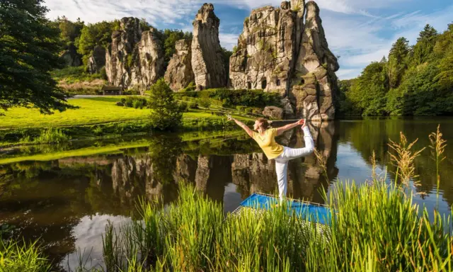 Frau praktiziert Yoga am See vor den Externsteinen NRW. Die Felsen spiegeln sich im Wasser.