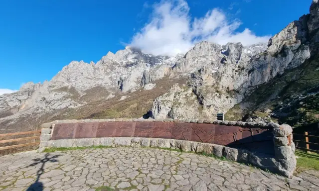 Impresionante mirador de los Picos de Europa con un muro de piedra y metal decorado. El sol ilumina las majestuosas montañas.
