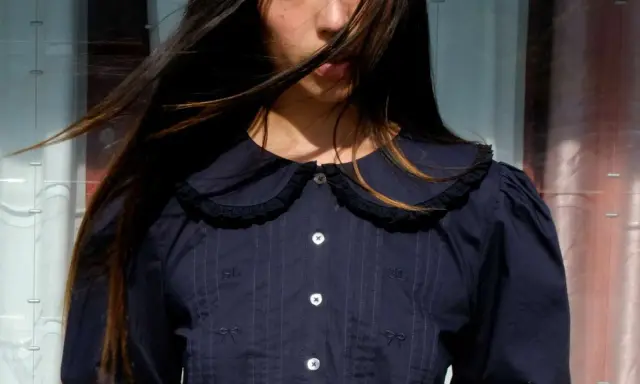 A young woman with windswept hair wears a dark blue blouse with a charming peter pan neckline, delicate embroidery, and puffed sleeves.