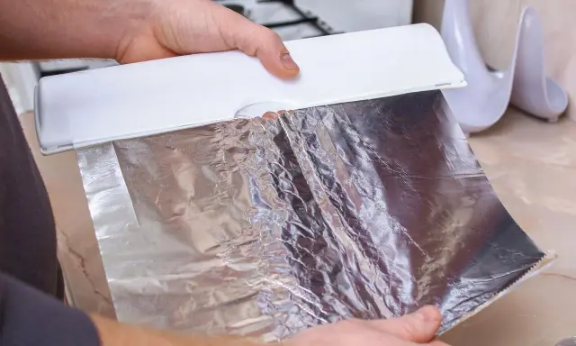 Ręce trzymają biały dozownik z folią aluminiową. Zastanawiasz się, która folia aluminiowa strona do pieczenia? Ta błyszcząca strona odbija ciepło.