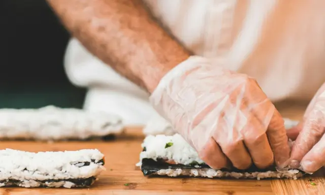 Hände mit Handschuhen rollen Sushi auf einem Holzbrett. So einfach ist Sushi selbst machen!
