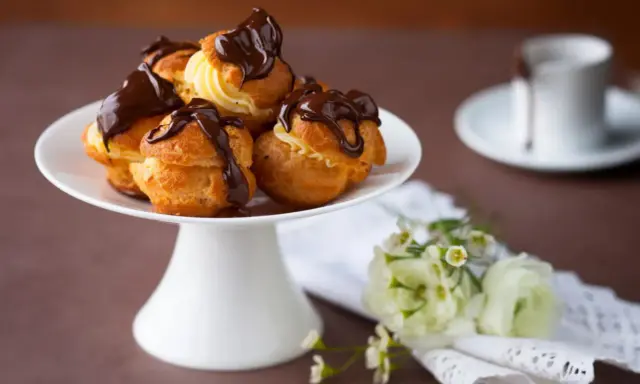Un plateau blanc présente des profiteroles gastronomiques garnies de crème et nappées de chocolat noir. Des fleurs délicates ajoutent une touche de fraîcheur.