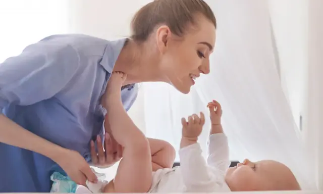 Uśmiechnięta mama zmienia pieluchę dziecku, zapewniając mu komfortowe szerokie pieluchowanie bioderek.