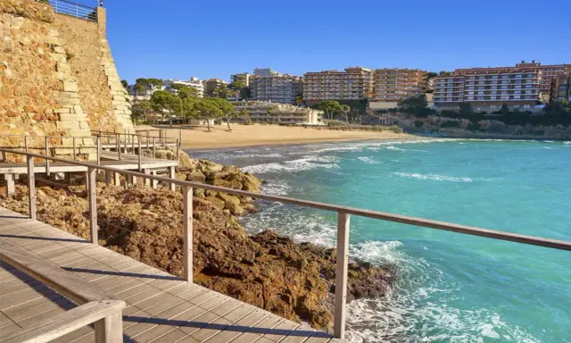 Paseo marítimo de madera junto a rocas y mar turquesa, con playa y edificios al fondo. Un tramo del camí de ronda Tarragona.