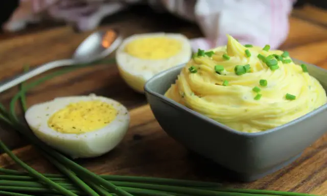 Cremige Sauce Tartar, garniert mit Schnittlauch, neben halbierten Eiern auf einem Holzbrett.