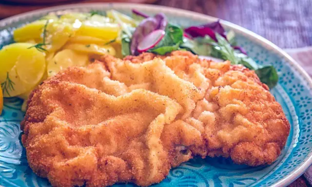 Ein knuspriges, originales Wiener Schnitzel auf einem blauen Teller, serviert mit Kartoffelsalat und frischem Grün.