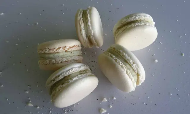 Cinq macarons vanille, délicatement posés sur une surface grise parsemée de miettes.
