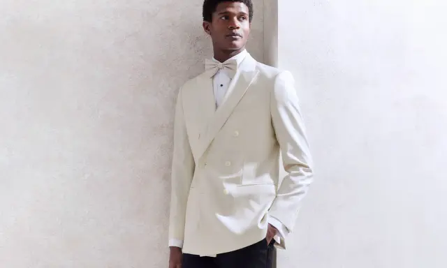 A Black tie wedding guest man in a cream double-breasted tuxedo with a matching bow tie, looking dapper.