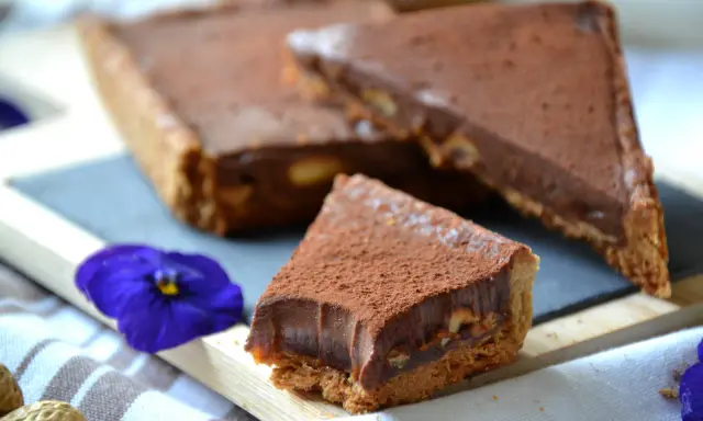 Tranche de tarte façon snickers, garnie de chocolat et de cacahuètes, saupoudrée de cacao. Une fleur bleue décore la présentation.