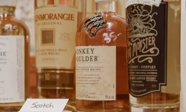 Flera flaskor whisky, inklusive Monkey Shoulder och Glenmorangie, står uppradade. En liten skylt med texten "Scotch" syns framför.