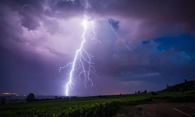 Un éclair zèbre le ciel violacé, illuminant un vignoble sous un orage de mer. La foudre frappe au loin, près d'une ville scintillante.