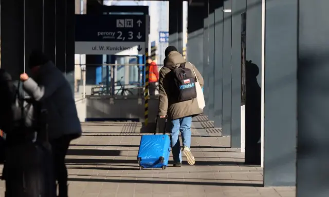 Osoba z niebieską walizką na kółkach i plecakiem idzie po peronie.