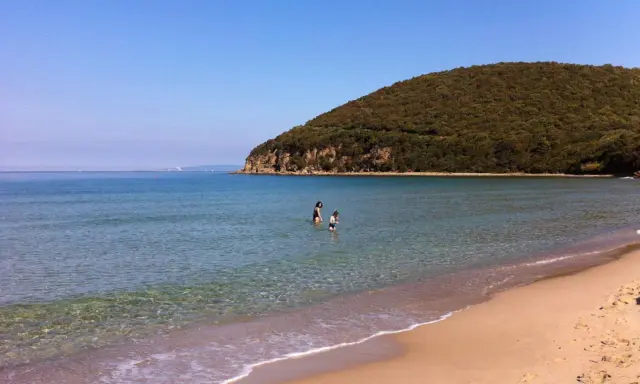 Spiaggia della Maremma con due bambini che giocano nell'acqua cristallina, sotto un cielo azzurro e una collina verdeggiante.