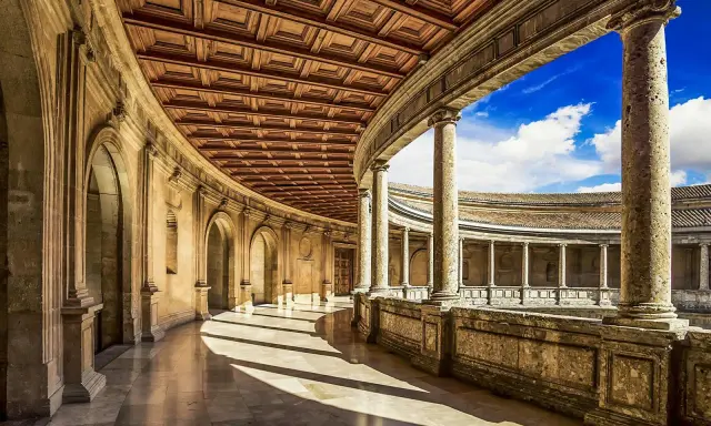 Ein sonnenbeschienener Arkadengang mit kunstvoller Holzdecke und Säulen, der zum Innenhof der Alhambra Moschee führt.