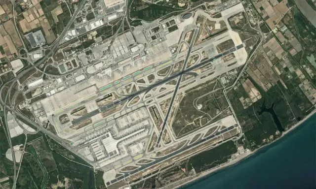 Widok satelitarny lotniska Barcelona El Prat. Widać pasy startowe, drogi kołowania, terminale i otaczające tereny rolnicze oraz morze.