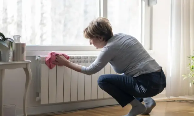 Mujer limpiando un radiador blanco con un paño rosa. Un ejemplo de cómo limpiar radiadores para mantener el hogar cálido y limpio.