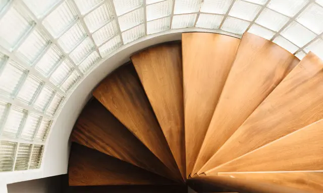 Escalera helicoidal de madera con peldaños que giran bajo un techo de bloques de vidrio translúcido.
