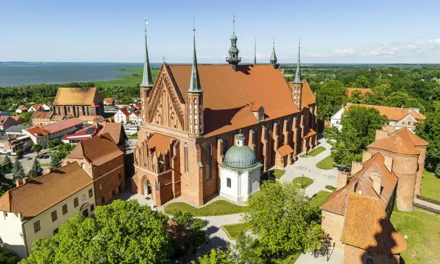 Zamek Frombork z katedrą i wieżami, otoczony zielenią, nad błękitną wodą.