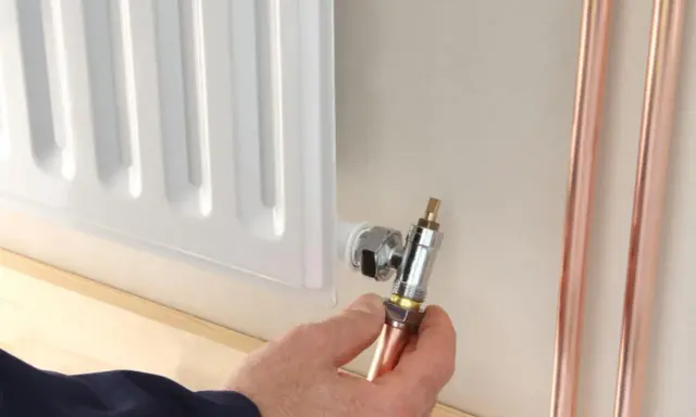 Instalator pokazuje, jak odkręcić dolny zawór grzejnika, trzymając miedzianą rurkę.