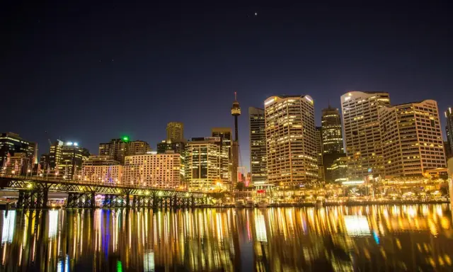 Sydney nocą, z mostem i odbijającymi się w wodzie światłami. Zastanawiasz się, ile się leci do Australii, patrząc na ten widok?