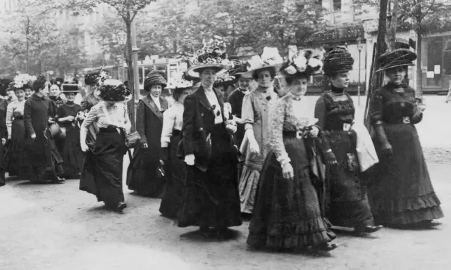 Frauen in langen Kleidern und Hüten marschieren entschlossen. Ein historisches Bild der feministischen Bewegung.